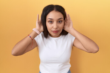 Young arab woman wearing casual white t shirt over yellow background trying to hear both hands on ear gesture, curious for gossip. hearing problem, deaf