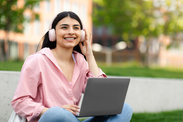 E-learning concept. Happy hispanic teen lady using laptop, wearing headphones listening to music, sitting in campus park