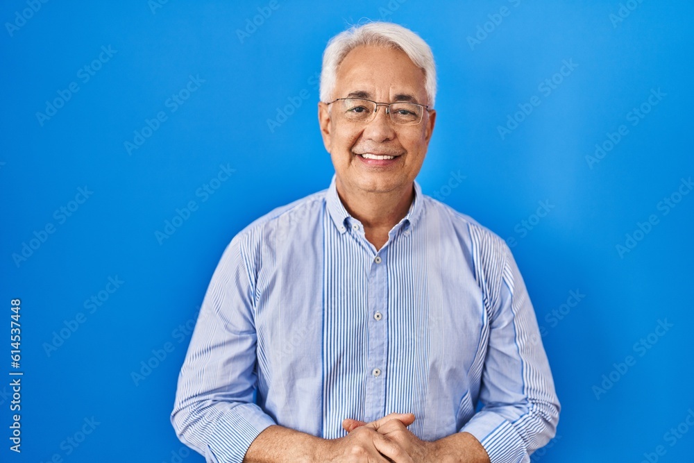 Wall mural Hispanic senior man wearing glasses with hands together and crossed fingers smiling relaxed and cheerful. success and optimistic