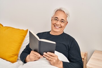 Senior man reading book sitting on bed at bedroom