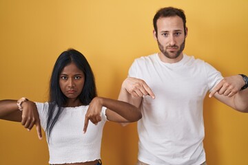 Interracial couple standing over yellow background pointing down looking sad and upset, indicating...