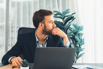 Businessman in black suit working on laptop at his workspace desk. Smart executive researching financial data and planning marketing strategy on corporate laptop at modern workplace. Entity