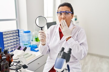 Middle age hispanic woman working at scientist laboratory holding magnifying glass covering mouth with hand, shocked and afraid for mistake. surprised expression