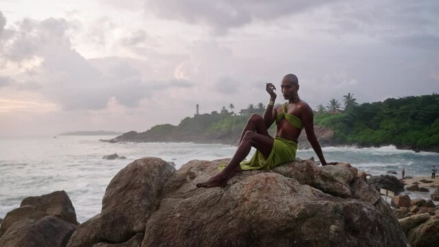 Non-binary black person poses gracefully sitting on top of rock in ocean at sunset slow motion. Trans ethnic fashion model in open tropical dress on peak by storm at dusk against sunrise