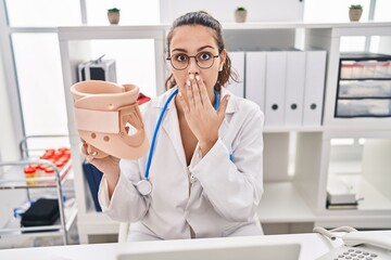 Young hispanic doctor woman holding cervical neck collar covering mouth with hand, shocked and...