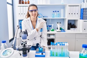 Young hispanic woman working at scientist laboratory cheerful with a smile of face pointing with hand and finger up to the side with happy and natural expression on face