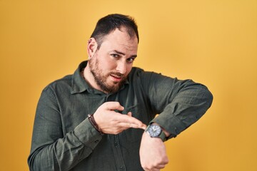 Plus size hispanic man with beard standing over yellow background in hurry pointing to watch time,...