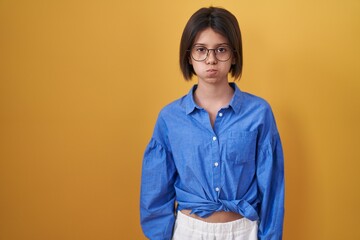 Young girl standing over yellow background puffing cheeks with funny face. mouth inflated with air,...
