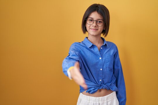Young girl standing over yellow background smiling friendly offering handshake as greeting and welcoming. successful business.