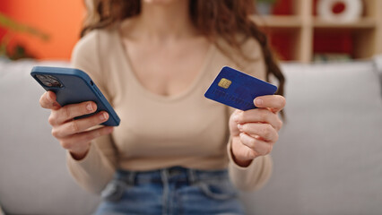 Young beautiful hispanic woman shopping with smartphone and credit card sitting on sofa at home