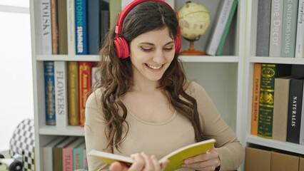 Young beautiful hispanic woman student listening to music reading book at library university