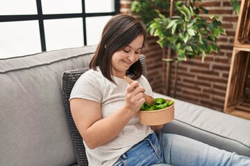Down syndrome woman eating salad sitting on sofa at home