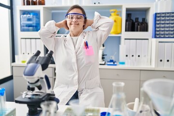 Hispanic girl with down syndrome working at scientist laboratory relaxing and stretching, arms and hands behind head and neck smiling happy