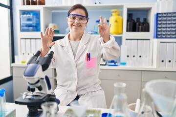 Hispanic girl with down syndrome working at scientist laboratory showing and pointing up with fingers number eight while smiling confident and happy.