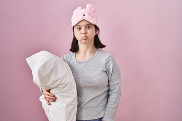 Woman with down syndrome wearing sleeping mask hugging pillow puffing cheeks with funny face. mouth inflated with air, crazy expression.