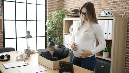 Young pregnant woman being fired packing belongings from workplace touching belly at office