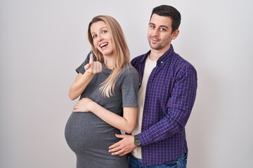 Young couple expecting a baby standing over white background beckoning come here gesture with hand inviting welcoming happy and smiling