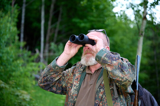 Gray-bearded senior hunter holding binoculars in hands and a rifle over his shoulder during hunting, walks through the forest, looks through binoculars and observes nature. Concept of hunt and travel