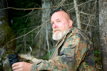 Gray-bearded senior hunter holding binoculars in hands and a rifle over his shoulder during hunting, walks through the forest, looks through binoculars and observes nature. Concept of hunt and travel