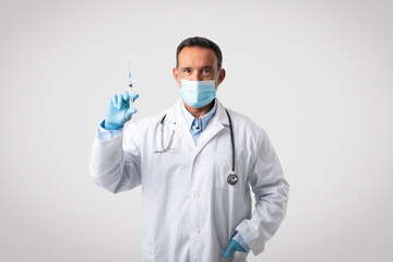 Serious confident caucasian mature doctor in white coat, protective mask and gloves holds syringe