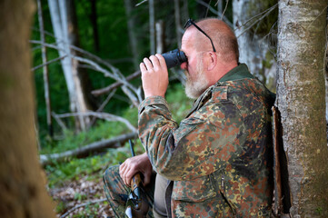 Gray-bearded senior hunter holding binoculars in hands and a rifle over his shoulder during hunting, walks through the forest, looks through binoculars and observes nature. Concept of hunt and travel