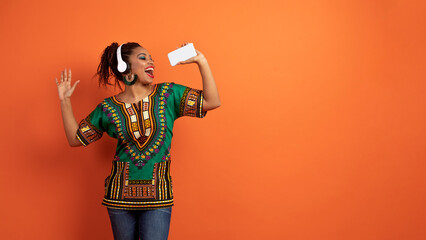 Happy black woman in african clothing singing, dancing on orange © Prostock-studio