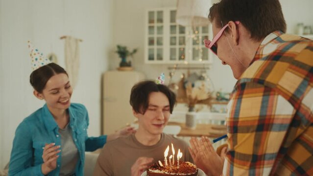 Portrait smiling birthday guy with cake in hands looks at burning candles makes a wish. Happy party with friends clapping hands and blowing horns on background. Celebration, festival mood concept.