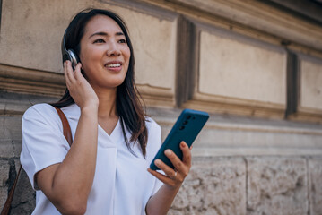 Young Japanese woman using headphone for listening nice music and walking in city. Girl happiness, smile face. Female love listen music that make her relax and enjoying life