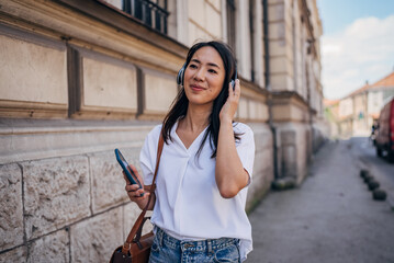 Young Japanese woman using headphone for listening nice music and walking in city. Girl happiness, smile face. Female love listen music that make her relax and enjoying life