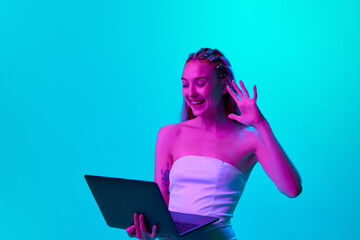 Portrait of beautiful young girl with stylish hairstyle talkign with video call on laptop against blue studio background in neon light. Concept of youth, emotions, beauty, lifestyle, ad