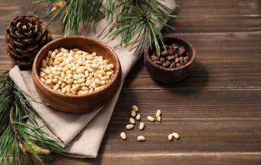 Obraz na płótnie Canvas Pine nuts in a bowl on napkin and a handful of unpeeled nuts on a brown wooden background with a branch of pine needles. Healthy diet snack.