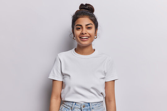 Horizontal shot of Indian woman with dark hair gathered in bun smiles pleasantly being in good mood dressed in casual t shirt and jeans isolated over white background. People and emotions concept