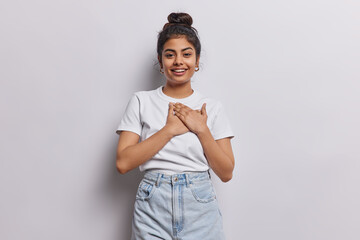 Indian woman dressed in casual white t shirt gently presses her hands to heart expressing deep...