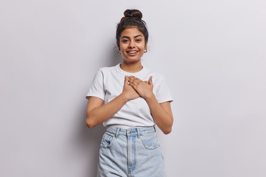 Indian woman dressed in casual white t shirt gently presses her hands to heart expressing deep appreciation and profound sense of being touched feels thankful dressed casually isolated on white wall