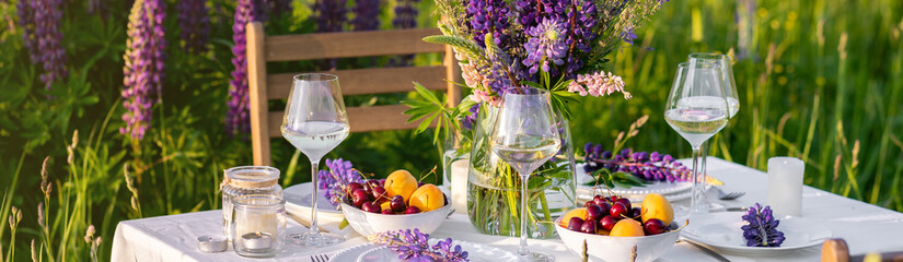 Beautiful countryside wedding of romantic dinner decor on blooming meadow with purple violet lupins flowers. Vase, wineglasses, cherry, apricot. Wooden table, basket. Sunset golden hour, summer banner
