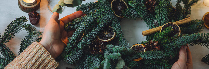 Young woman in knitted sweater doing handmade Christmas wreath from natural materials: fir tree branches, dry citrus slices, cones, cinnamon sticks. Wooden table, cozy light, home atmosphere. Banner