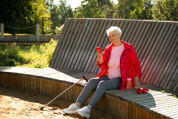 An elderly woman with short gray hair sits on a wooden bench in the park and watches the news on a smartphone