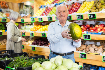 European senior man standing in salesroom of greengrocer with melon in hands. Woman choosing fruits...
