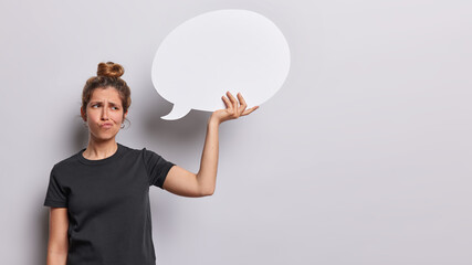 Displeased young woman has dissatisfied expression dressed in casual black t shirt holds communication bubble isolated over white background copy space for your advertisement. Place your promo here