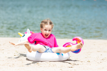 Adorable happy smiling little girl in pink t-shirt has fun with swimming ring unicorn on beach...