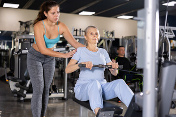 Female fitness trainer is engaged explains how to exercise on the simulator to an elderly woman in the gym