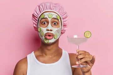 Impressed adult man holding refreshing cocktail gently applies facial clay mask accompanied by...
