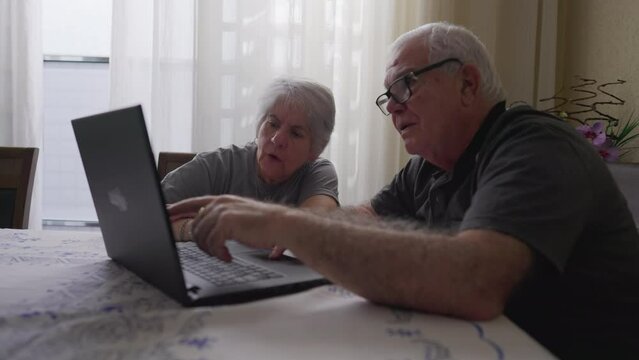 Elderly couple in front of laptop sitting in living-room at home. Senior husband pointing at computer screen showing content online to wife