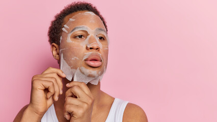 Serious African guy enjoying moisturizing procedure applies beauty sheet mask on face concentrated...
