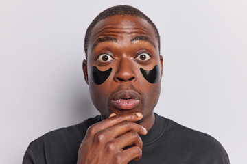 Shocked dark skinned man keeps hand on chin stares bugged eyes at camera applies beauty patches to reduce wrinkles and puffiness wears black t shirt isolated over white background. Studio shot