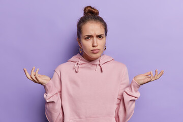 Horizontal shot of displeased young European woman gazes at camera with questioning look her brows furrowed in bewilderment dressed in casual pink sweatshirt isolated over purple background.