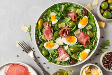 Prosciutto salad with parmesan, olives, eggs and arugula in a plate on gray background. Italian food. top view