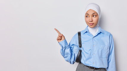 Waist up shot of serious young Muslim woman dressed formally carries small bag on shoulder points away on blank space for your advertisement isolated over white background. Click on this link