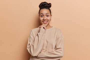 Fototapeta premium Waist up shot of cheerful Latin woman keeps hand on chin smiles gladfully has curious expression dressed in casual pullover isolated over brown background. Optimistic millennial female model