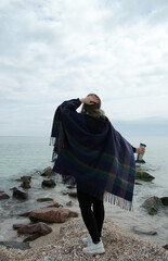 Woman in a poncho on the seashore in warm clothes and with a cup of coffee, picnic, spring on the beach